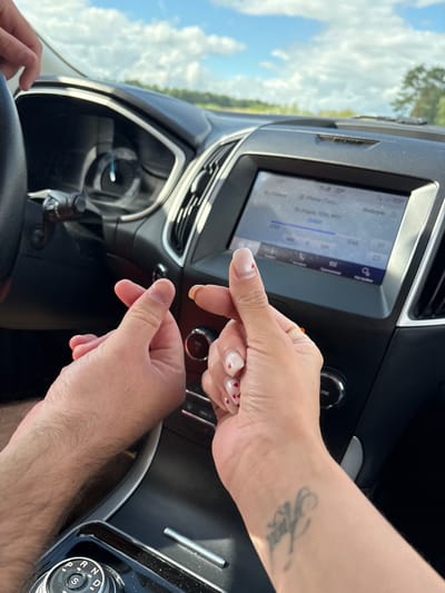 The image shows a close-up view of two people's hands inside a car. The left hand, presumably belonging to a male due to its size and hairy appearance, is making a "finger heart" gesture. The right hand, belonging to a female, is also making a similar gesture, with manicured nails decorated with small red hearts. The background reveals the interior of a modern car, including a dashboard with a navigation screen displaying a radio interface and a gear shift knob labeled "P R N D S". The overall setting appears to be a sunny day, as indicated by the light streaming through the car's windshield and the partially visible trees and sky outside. The image does not provide any specific visual cues that would allow for identification of the location within Мазалаўскі сельскі Савет, Belarus.