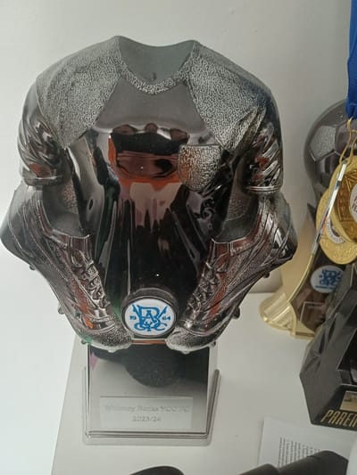 The image shows a trophy shaped like a football boot, displayed on a white surface. The boot is predominantly black with silver accents and has a textured finish on the sides. A blue and white circular emblem is visible on the side of the boot, featuring the numbers "19" and "64" alongside a stylized blue logo. The trophy sits on a clear acrylic base, which has text etched onto it. The visible text on the base reads "Whinney Banks YOO FO" and "2023/24".
In the background, other awards and medals are partially visible. One medal is gold and has text on it, possibly relating to football. Another award is gold and black, with "PAREN" visible on its side. A blue ribbon is also seen, attached to one of the medals.
The scene appears to be a display of football awards, possibly in a trophy cabinet or on a shelf. The lighting is bright, suggesting it might be daytime or indoors with good lighting. There are no people visible in the image. The overall impression is one of celebration and recognition for achievements in football.