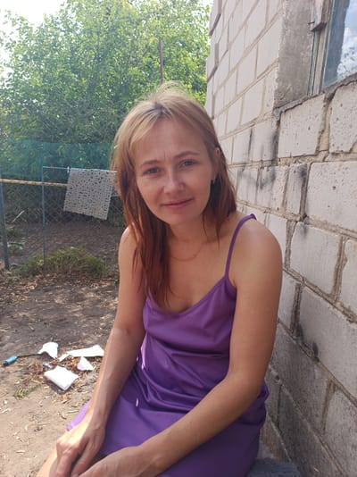 A fair-skinned Caucasian woman with shoulder-length, reddish-blonde hair and blue eyes is seated in an outdoor environment. She is wearing a sleeveless purple dress and a thin gold necklace. The woman is positioned in front of a light-colored brick wall, with a chain-link fence and dense green foliage visible behind her to the left. The ground is covered with dirt, scattered leaves, and some white debris. The scene appears to be in a rural or semi-urban setting, potentially in Saksahan, Ukraine, given the architectural style and vegetation.