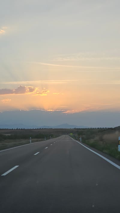 This media file depicts a view of a road stretching towards a horizon at sunset. The road itself is dark asphalt with white lane markings and a white edge line. To the left of the road, the sky is filled with soft, diffused light, transitioning from a pale blue to warm oranges and yellows as the sun sets. Wispy clouds are scattered across the sky, some illuminated by the setting sun. In the distance, a range of mountains or hills is visible, appearing as dark silhouettes against the colorful sky. The overall scene suggests an open, rural landscape. The time of day is clearly sunset due to the warm, golden light and the angle of the sun. The weather appears clear with some high-altitude clouds. There is a road sign with yellow and black markings on the right side of the road, which is partially obscured by the grass and vegetation bordering the road. The image conveys a sense of calm and transition as day turns into night.