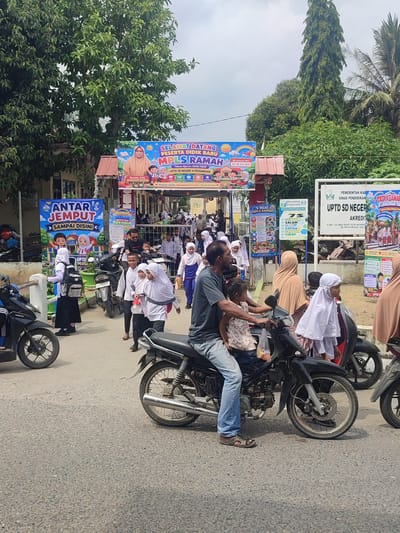 The image depicts an outdoor scene at what appears to be the entrance of a school, likely UPTD SD NEGERI 4 PEUSANGAN in Panton Rah, Indonesia, given the signage. A crowd of people, primarily children in school uniforms, are gathered on a paved road. Many of the children are wearing white shirts, black skirts or trousers, and hijabs, suggesting a religious or cultural context.

Several motorcycles are parked or being ridden, with some individuals on them. One prominent motorcycle in the foreground carries a man wearing a dark t-shirt and blue jeans, with a young girl in a pink and white patterned top on his lap. Another motorcycle is ridden by a man in a black t-shirt and blue jeans. Children are seen walking and standing around, some carrying backpacks.

Large banners are displayed, including one that reads "SELAMAT DATANG PESERTA DIDIK BARU MPLS RAMAH" and another with "ANTAR JEMPUT SAMPAI DISINI." These banners indicate a welcome event for new students, likely related to the "Masa Pengenalan Lingkungan Sekolah" (MPLS), or school environment introduction period. The overall atmosphere appears to be busy and celebratory, with the school day or an event just beginning or concluding.

The environment includes a building structure with a red roof visible behind some of the banners, and lush green trees and palm trees line the background, suggesting a tropical or subtropical climate. The sky is partly cloudy. The road surface is asphalt with visible aggregate. The time of day appears to be daytime, with moderate sunlight casting shadows.