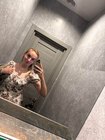 A woman with blonde hair is taking a mirror selfie in a modern restroom. She is wearing a white floral print dress with puff sleeves and a necklace. In her right hand, she holds a smartphone with a gold case, and her left hand is resting on her bag. The restroom features large grey tiles on the walls and floor, a wooden door, and a circular ventilation grate on the wall. The lighting suggests it is daytime. The woman has a slight smile and appears to be in a casual setting.