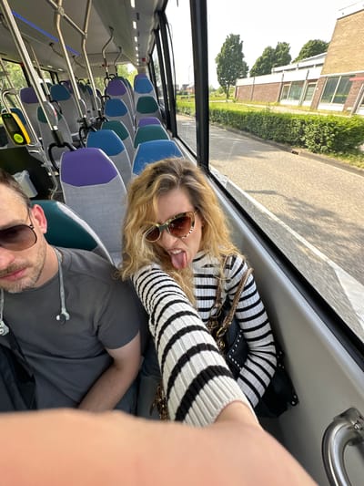 Inside a bus in Dronten, Netherlands, a woman with blond hair and sunglasses, wearing a black and white striped sweater, sticks her tongue out towards the camera. Beside her, a man with sunglasses and a gray shirt is visible. The interior of the bus features rows of seats with alternating blue and purple headrests. Outside the bus window, buildings, trees, and a street are visible, suggesting a cityscape.