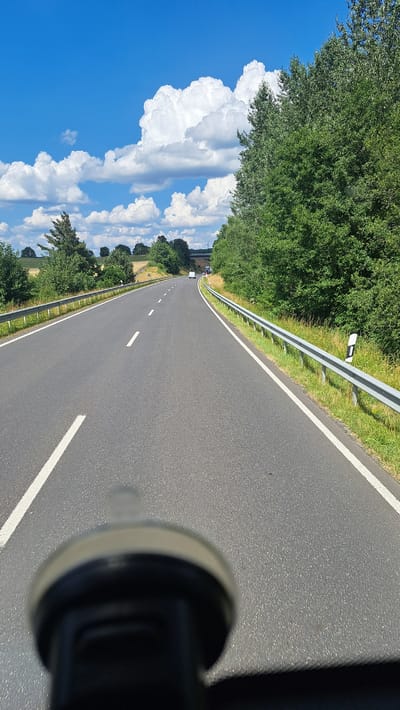 A first-person perspective shot captures a view down a two-lane asphalt road with a white dashed line marking the center and solid white lines along the edges. The road curves gently to the left and disappears over a rise in the distance. To the right of the road, a metal guardrail runs along the edge, bordered by a grassy shoulder and dense green trees. To the left of the road, a similar grassy shoulder transitions to fields and scattered trees. The sky above is a vibrant blue with large, fluffy white cumulus clouds. In the far distance, a white vehicle is visible on the road, and a bridge or overpass is suggested. The foreground is blurred and occupied by a dark, round object, possibly a car mount for a camera or phone. The scene is set in a rural environment, specifically identified as being near Rennerod, Germany. The time of day appears to be midday, given the bright sunlight and clear sky.