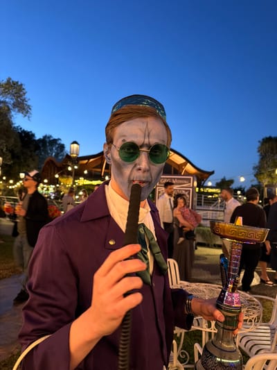 This full-shot, portrait-oriented image captures an outdoor evening social event, likely a wedding, as indicated by the visible "GRA WEDDING" sign in the background. The sky is a deep twilight blue, illuminated by numerous string lights and other decorative lighting fixtures that create a festive atmosphere.
The primary subject is a person in a theatrical costume, prominently featured in the foreground. Their face is painted a light blue or grey with dark, skeletal-like contouring under the eyes and on the cheeks. They wear round, green-tinted sunglasses and a patterned blue-green skullcap or head covering. The individual is dressed in a dark purple jacket over a white shirt, with a green tie, and holds an ornate, multi-colored hookah. Their right hand holds the black hose of the hookah to their mouth, while their left hand supports the shimmering base and stem of the hookah, which features metallic gold accents and intricate patterns.
In the blurred background, several casually dressed people are visible, some standing and others seated on white, ornate outdoor chairs around tables. A distinctive wooden structure with a curved roof is also seen behind the main subject. The scene is set outdoors with visible trees and hints of greenery, contributing to a lively and relaxed social gathering under the evening sky.