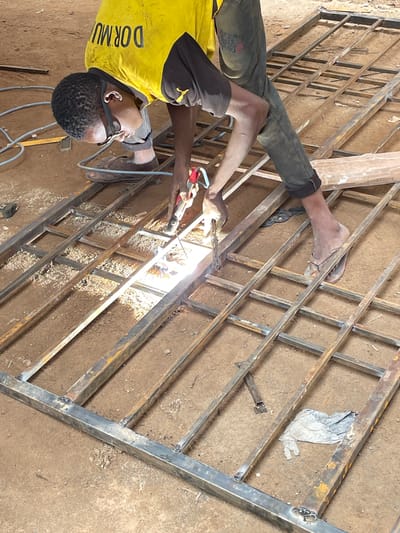 In Okinano, Nigeria, a young man is seen welding a metal gate outdoors. He is wearing a yellow t-shirt with black sleeves and black pants, and he has safety glasses on. The welding is taking place on a flat, dirt surface, which is typical of many outdoor work areas in the region. The metal gate is laid out on the ground, and the man is working diligently to join the metal bars together. Sparks are flying from the welding torch, illuminating the immediate area and highlighting the intense heat of the process. The surrounding environment appears to be a workshop or a construction site, with other metal materials and tools potentially present but not clearly visible in the frame. The overall scene depicts a craftsman at work, contributing to the infrastructure or livelihood of the community.