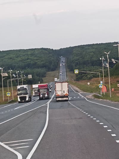 The image shows a view of a highway in the city of Kologreyevka, Russia. The road is paved and has multiple lanes, with white dashed lines indicating the lanes. Several large trucks are on the highway, traveling in the same direction. Some of the trucks are white, while one is red. There are also smaller vehicles visible in the distance. The highway is lined with trees on either side, forming a dense forest. The sky is overcast with a light gray color. There are streetlights and road signs along the side of the road, including pedestrian crossing signs. The overall scene depicts a typical highway environment in a rural or semi-rural area.