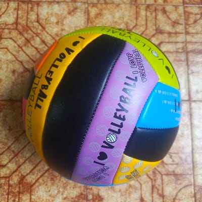 A multi-colored volleyball lies on a brown, tiled surface. The volleyball has sections of yellow, orange, pink, black, purple, and blue. Text and volleyball icons are printed on several sections of the ball. The yellow section has the word "VOLLEYBALL" printed in black. The purple section has the words "I LOVE VOLLEYBALL" and many small volleyball icons. The blue section has the words "I LOVE VOLLEYBALL" repeated several times, along with heart icons. The background surface appears to be an outdoor tiled patio or walkway. There are no people or any other objects visible in the image. The scene is static and still. The location of Los Teques, Venezuela is not discernible from the image content itself.