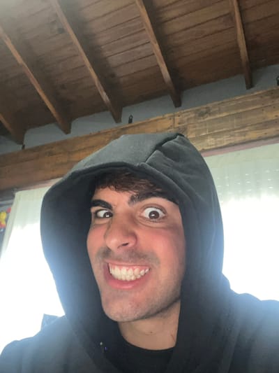 A close-up selfie captured in Sarandí, Argentina, features a person, likely male, with a playful and exaggerated expression. The subject is wearing a dark hooded sweatshirt, with the hood up, framing their face. They have dark hair, dark eyebrows, and wide, brown eyes that are looking upwards and slightly to the right. A broad, open-mouthed smile reveals white teeth, suggesting a moment of amusement, surprise, or excitement.

The background on the left side of the frame shows a rustic indoor setting with warm-toned wooden planks and exposed wooden beams, arranged diagonally due to the image's tilt. On the right and bottom, a large area of bright white indicates a strong light source, possibly a window, which creates a high-contrast lighting effect. The overall image is rotated, giving it a dynamic and informal feel. There is no visible text.