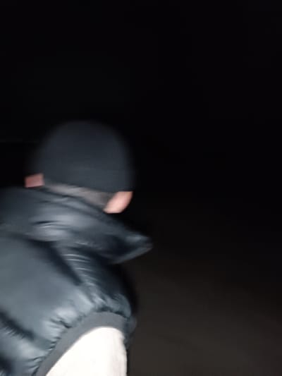 The image displays a single individual within a profoundly dark, low-light environment in Shamsikul, Uzbekistan. The subject is positioned with their back and right profile towards the viewer, exhibiting significant motion blur consistent with movement. They are attired in a dark beanie, a dark, likely black, quilted vest, and a lighter-toned garment visible beneath the vest. Only an indistinct outline of the right side of the head and a partial, light-skinned cheek area are discernible. The background is featureless and appears entirely black, with subtle, dark, indistinct ground visible beneath the subject. A faint, small, reddish-orange rectangular object or artifact is barely perceptible in the lower-left background. No other individuals, objects, or specific interactions are evident within the frame.