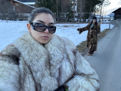Two individuals are depicted in a snowy, mountainous landscape in Saanen, Switzerland.
In the foreground, a female is shown from the chest up, positioned for a selfie. She wears large, dark, wrap-around sunglasses and small silver hoop earrings, with her dark hair pulled back. Her lips are slightly pursed, and she looks directly at the camera. She is clad in a thick, light-colored fur coat exhibiting shades of silver, grey, and brown.
Approximately ten meters behind her, to the right of the frame, a second female stands on a paved road. She wears a long, dark brown fur coat, dark pants, light brown Ugg-style boots, a black beanie, and small white-framed sunglasses. She extends her right arm forward and to her right, pointing with her index finger, while holding a light-colored smartphone vertically in her left hand.
The environment includes a paved road running from the foreground into the distance, bordered on the left by snow-covered ground interspersed with brown grass and a low white wire fence. Numerous pine trees and other bare trees are visible in the background. Two traditional wooden structures, one a multi-story chalet and another partially visible, are present among the trees. Distant snow-capped mountains are discernible under a daylight sky, suggesting winter conditions.