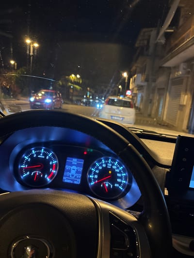 The image is taken from inside a vehicle, focusing on the dashboard and steering wheel, with a nighttime urban street scene visible through the windshield. The dashboard instrumentation is lit, displaying data such as speed in km/h and tire pressure (TPMS). The exterior view shows streetlights, buildings, and other vehicles, indicating the location as the city of Polistena, Italy.