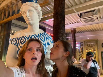 In Solnechnoye, Russia, two young women pose for a selfie with a large, white statue of a woman wearing a blue and white patterned garment and a blue crown. The statue is positioned in the foreground, behind the two women. The woman on the left has her mouth open in a surprised expression and is looking at the camera. The woman on the right is looking upwards, towards the statue's head. Behind them, a tree trunk rises towards a ceiling with exposed beams and an air conditioning unit. Further in the background, several people are visible, including a man wearing a baseball cap who appears to be adjusting his clothing. The setting appears to be an indoor establishment, possibly a restaurant or cafe, with decorative elements and lighting suggesting a public space.