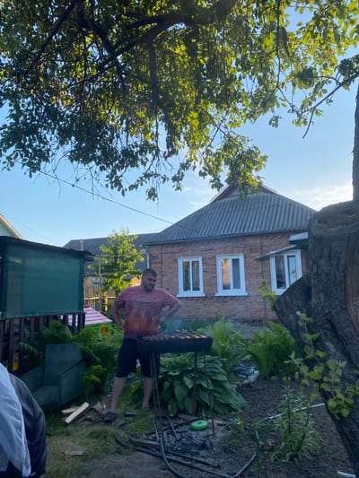 An outdoor scene, likely a backyard in Romny, Ukraine, captures a man standing by a barbecue grill in what appears to be a rural or suburban setting. The man, wearing a pink t-shirt and dark shorts, is positioned with his hands on his hips, facing the grill. The grill contains meat on skewers, suggesting food is being cooked.

The environment is lush with greenery, featuring large hosta plants, ferns, and a prominent tree with branches extending over the scene, dappling it with sunlight. In the background, a brick house with a corrugated roof and white-framed windows stands under a clear blue sky. Adjacent to the house is another structure, possibly a shed or outbuilding, covered with a green tarp.

The ground is a mix of grass and soil, with scattered pieces of wood, a green circular object, and other debris suggesting recent activity or ongoing yard work. The overall impression is one of a casual, domestic gathering or preparation for one, set against a backdrop of typical Ukrainian countryside architecture.