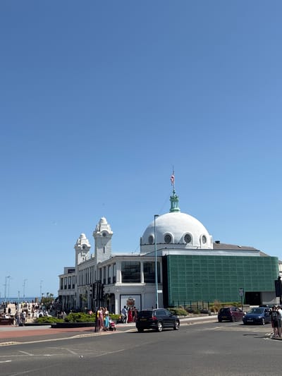 The image shows the Spanish City building in Whitley Bay, North Tyneside, United Kingdom. The white dome and architectural details of the building are visible against a clear blue sky. A road runs along the right side of the image, with cars parked and people walking along the pavement. There are a few trees and benches. In the background, the sea is visible.