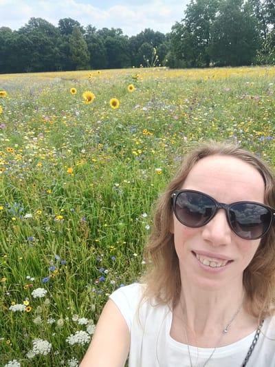 The image depicts a woman in the foreground, smiling and wearing sunglasses, with a vast field of wildflowers and sunflowers extending behind her. The field is a vibrant mix of yellow, purple, and white flowers, with tall green grass interspersed. In the background, a dense line of trees marks the edge of the field, and a cloudy sky is visible above. The scene appears to be an outdoor natural setting, likely a meadow or park. Based on the provided hints, the location is specified as Isselburg, Germany.