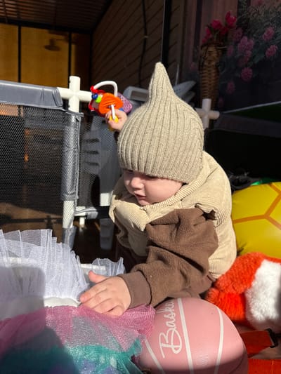 A baby wearing a brown and beige outfit and a pointed knit hat is positioned head-down inside or over a portable playpen. The baby is holding a brightly colored toy with one hand while the other hand touches a multi-colored tulle ruffle. The playpen has a white frame and a black mesh side. The background includes toys, a basket of flowers, and other items, suggesting the scene may be indoors or in a contained outdoor space. The location is Кипчак, Russia, but without further information, it is impossible to pinpoint a specific area within the city.