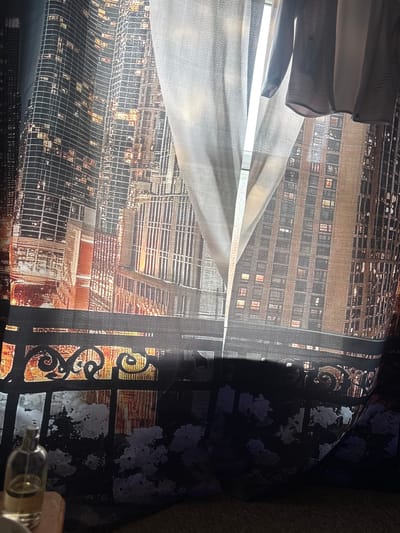 The image depicts a window with curtains printed with a nighttime cityscape. The left curtain shows tall buildings with illuminated windows, suggesting a city like Mansfield. A sheer white curtain is drawn partly across the window, revealing more of the cityscape behind it. Hanging from the top of the window, in front of the sheer curtain, are two items of clothing, possibly a shirt and a skirt, made of a light-colored fabric. A dark, ornate railing is visible along the bottom of the printed curtain. In the lower left corner of the image, a clear glass bottle with a spray nozzle is partially visible, resting on an orange surface. The overall scene suggests an indoor setting, looking out through a window at a city scene printed on the curtains.