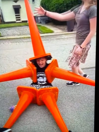 In an outdoor, paved urban environment, a person is shown at ground level, positioned in the center of the frame, adorned with an arrangement of orange traffic cones. The cones are strategically placed to encase the individual, with some forming a structure around their torso and head, and others extending outwards like limbs. The person appears to be wearing a black t-shirt with a graphic design visible on it.
Standing behind and to the right of the cone-encased individual is another person, a woman, whose left arm is extended towards the person in cones, palm open in a gesturing motion. She is wearing a grey t-shirt and a patterned skirt, with black athletic shoes. The background shows a residential building with a porch and door, a manicured lawn, and dense green shrubbery, typical of a suburban or urban streetscape. The ground is asphalt or concrete, showing some texture and minor debris. The overall lighting suggests daytime.