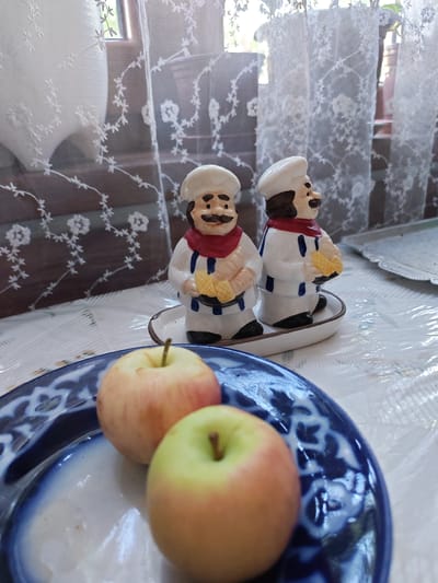 In Kogon City, Uzbekistan, a still life composition unfolds on a table covered with a white plastic tablecloth and a patterned cloth underneath. Two ceramic figurines, dressed as chefs with white hats, red scarves, and white uniforms with blue stripes, stand on a small white oval dish. Each chef holds a yellow object, possibly corncobs. In the foreground, two apples, one predominantly red and the other with yellow and red hues, rest on a blue and white patterned plate. The background features a window with white lace curtains and wooden blinds, allowing diffused natural light to illuminate the scene.
