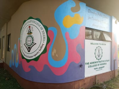 The image captures the exterior of the Administrative Staff College of Nigeria (ASCON) in Topo-Badagry, Nigeria. The building's wall is adorned with the college's emblem and name. The emblem is prominently displayed, surrounded by the words "THE ADMINISTRATIVE STAFF COLLEGE OF NIGERIA - the natural place for human capacity building". Adjacent to the emblem, a colorful abstract design adorns the wall. Nearby, a white section of the wall bears the inscription "WELCOME TO ASCON" alongside the college's emblem and the institution's name. Below, "TOPO-BADAGRY Founded in 1972" is written in green. Above the "WELCOME" sign, the words "Elevating Public Service for Operational Excellence through training" are inscribed on the wall. The base of the wall is painted brown, with greenery visible at the foot of the building.