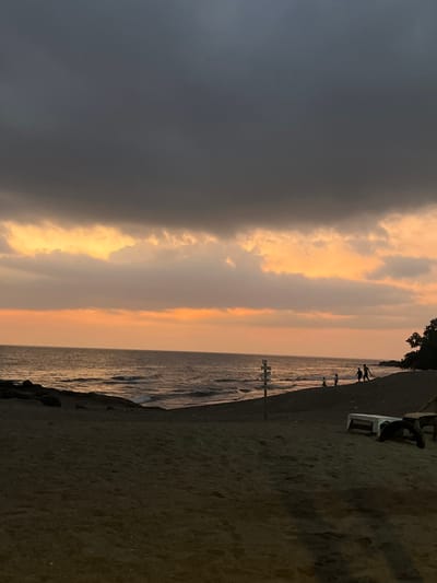 The image depicts a coastal scene at dusk or sunset in Desa Batu Layar Barat, Indonesia. The upper two-thirds of the frame are dominated by a heavy, dark grey cloud cover, beneath which a horizontal band of vibrant orange and yellow light from the setting sun is visible along the horizon. Below the horizon, the ocean surface is dark with visible waves breaking onto a sandy beach. To the left, a dark, rocky outcrop extends into the water. In the center-right, a multi-directional signpost with multiple white, arrow-shaped signs is planted in the sand near the waterline. Further right along the sandy beach, a group of approximately four to five indistinct human silhouettes are visible, appearing to walk or stand near the water's edge. Dark tree foliage is present on the far right edge of the frame, bordering the beach. In the lower right foreground, two dark, low-lying structures, possibly wooden benches or tables, are partially visible on the sand. The overall lighting is low, emphasizing silhouettes and the orange glow of the sunset.