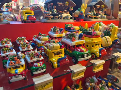 The image depicts a detailed display of ceramic souvenir figurines, likely within a retail shop in Girón, Colombia. The foreground and midground feature multiple rows of miniature vehicles, primarily "chivas" (traditional Colombian rural buses) and cars, along with some donkey figures. These figurines are brightly colored, predominantly yellow, red, green, and blue, and are depicted as overloaded with small, sculpted items such as fruits, baskets, and luggage, typical of rural transportation. Many of the vehicle figurines have "COLOMBIA" inscribed on their side or front. They are arranged on a transparent glass shelf, which rests on a red shelf or display unit. Several small, light-colored wooden blocks serve as price tags beneath some of the figurines; one visible tag displays "COP 34,900" and "1 USD 23999". In the background, on a higher red shelf, more elaborate ceramic displays are visible, including a miniature rural scene with figures under a thatched hut and a large yellow donkey figure. No people are present in the scene, and there are no discernible actions or interactions occurring. The environment suggests an indoor, well-lit commercial space, possibly a gift shop specializing in Colombian handicrafts.