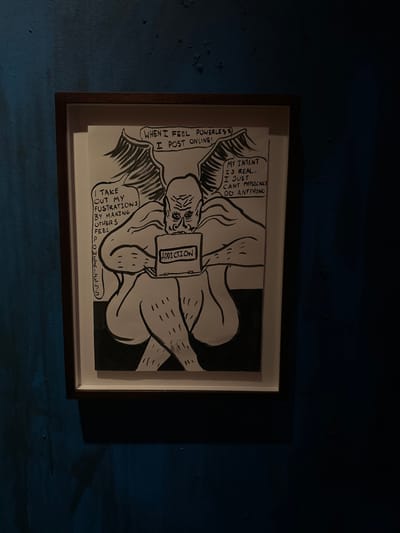 A framed black-and-white ink drawing is centrally displayed on a textured, dark teal wall in a dimly lit interior space within the City of Westminster, United Kingdom. The artwork depicts a large, muscular, bald, winged figure with a prominent nose and furrowed brow, sitting cross-legged. The figure holds a rectangular device, possibly a laptop or tablet, emblazoned with the word "ADDICTION" in block capitals. Three speech bubbles emanate from the figure: one above reads "WHEN I FEEL POWERLESS I POST ONLINE!", another to the left states "I TAKE OUT MY FRUSTRATIONS BY MAKING OTHERS FEEL POWERLESS", and a third to the right declares "MY INTENT IS REAL... I JUST CAN'T PHYSICALLY DO ANYTHING". The drawing is rendered in a bold, illustrative style with thick outlines and minimal shading. It is housed within a dark brown frame with a white inner mat. The wall surface appears uneven and slightly distressed, suggesting a painted or rendered finish. The low ambient lighting casts a faint shadow beneath the framed artwork. The exact location within the City of Westminster is not identifiable from the visual evidence.