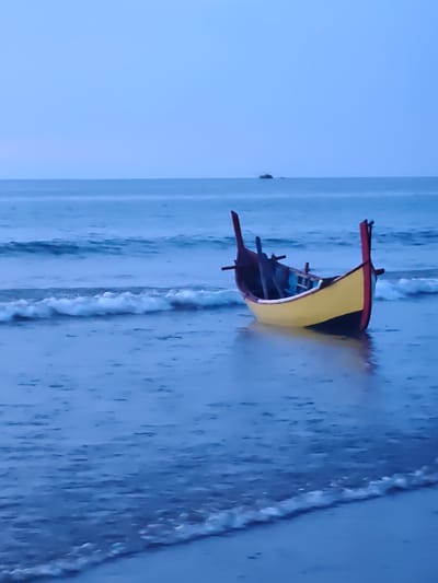 A yellow and red fishing boat rests on the shallow, wet sand at the edge of a calm ocean. The water reflects the overcast sky, which transitions from a pale blue at the horizon to a deeper indigo overhead. Small waves lap gently against the shore, creating a foamy white line where they meet the sand. The boat appears empty, with its dark interior contrasting with its bright exterior. The scene is serene and quiet, suggesting either dawn or dusk. There are no people visible in the image. This scene is located in Curehtunong, Indonesia.