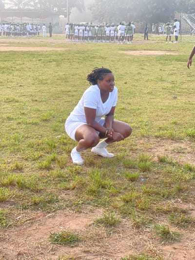 The image depicts an outdoor scene at a parade ground in Emure-Ekiti, Nigeria. In the foreground, a young Black woman is squatting on the grass, wearing a white t-shirt and white shorts, with white socks and sneakers. She has braided hair and is looking to her right. In the background, a large group of young people, appearing to be in a uniformed assembly or parade, stand in formation on a grassy field. Some are wearing white shirts and green shorts, while others are in white. Further in the background, there are structures that suggest permanent buildings and possibly tents, along with a goalpost on the right side of the frame. The overall environment appears to be a dusty, open area with sparse vegetation and a hazy atmosphere, indicative of an outdoor training or assembly ground.