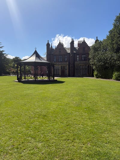 The image depicts a scenic view in Warrington, United Kingdom, featuring a well-maintained lawn, a gazebo, and a large building in the background. The grass is lush and green, covering a significant portion of the frame. A gazebo with a dark frame and a light-colored canopy stands to the left of the lawn. Behind the gazebo, a substantial building with multiple levels and intricate architectural details is visible. The building is constructed with dark brick or stone, featuring numerous windows and decorative elements on its roofline. The sky is a clear blue with scattered white clouds, providing a bright backdrop for the scene. The overall impression is one of a tranquil and well-kept public park or garden within the city of Warrington.