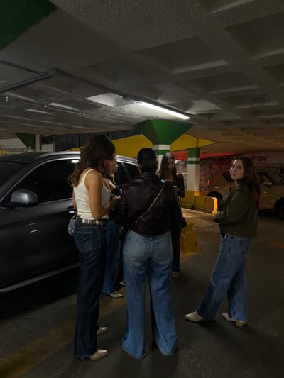 The scene depicts five females gathered within an illuminated section of a concrete parking garage in Guanajuato City, Mexico.

**Environment:** The overhead concrete ceiling features recessed square panels and exposed conduits, with multiple fluorescent light fixtures providing illumination. Large pillars, painted with green and white horizontal stripes, punctuate the space. One pillar also exhibits a yellow section. The ground is paved, possibly asphalt, with faded yellow markings. A dark grey SUV is parked on the left, partially obscuring the leftmost individual. In the far background, a lighter-colored vehicle is partially visible near a stone wall, which shows a reddish glow.

**Individuals:**
1.  **Leftmost:** Partially visible from behind, presenting long dark hair and a dark top.
2.  **Second from left:** Standing with her back to the camera, she wears a white tank top, dark blue jeans, a metallic silver belt, and gold-toned shoes. A light-colored shoulder bag with a distinct pattern is slung across her body.
3.  **Center:** Positioned with her back to the camera, she wears a dark reddish-brown leather-like jacket, light blue wide-leg jeans, and gold-toned shoes or boots. A delicate gold chain or strap crosses her upper back.
4.  **Fourth from left:** Mostly obscured by the person in the center, she has long dark hair and wears dark attire.
5.  **Rightmost:** Facing the camera, she has long brown hair and wears a dark olive-green jacket, blue jeans, and white pointed-toe boots with a low heel. She looks directly towards the camera with a neutral expression.

**Actions/Interactions:** The individuals appear to be conversing or waiting, standing in close proximity to one another. Three individuals face away from the camera, seemingly engaged with each other, while the individual on the far right directly observes the camera. No overt physical interactions are observed.
