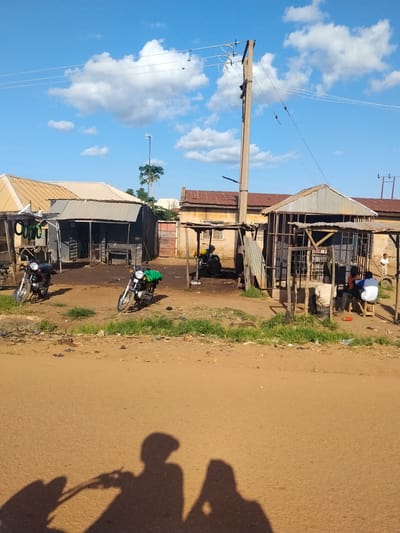 This image depicts a daytime scene in a dusty, outdoor environment, likely in Mangu Town, Nigeria. The sky is a vibrant blue with scattered white clouds, suggesting fair weather. The foreground is dominated by a sandy, reddish-brown road where the shadows of two people on a motorcycle are cast, indicating the photographer's perspective.
Behind the road, a collection of small, open-fronted structures and buildings line the area. These appear to be local shops or stalls, constructed from corrugated metal and wood, with some showing signs of wear. Several motorcycles are parked in front of these establishments, suggesting their importance for transportation or commerce in this location.
In the mid-ground, a group of people are gathered near one of the structures. Some are seated, appearing to be engaged in conversation or resting. One person stands slightly apart, looking towards the buildings. The overall atmosphere seems casual and perhaps a bit sluggish, typical of a warm, bright day in a bustling, informal marketplace.
A prominent utility pole with electrical wires stretches across the upper portion of the image, a common sight in many populated areas. The scene is characterized by its unpaved ground, modest architecture, and the presence of everyday objects like motorcycles and makeshift stalls, all contributing to a strong sense of place in a rural or semi-urban Nigerian setting. There is no visible text in the image.