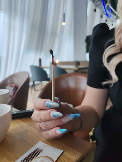 The image shows a close-up of a person's hand holding a lit match. The match has a red and orange flame and is burning down. The person's nails are long and painted with a blue and gold design. In the background, there are chairs and tables, suggesting an indoor setting like a cafe or restaurant. The overall ambiance is casual. There is no information about the location being Amursk, Russia, in the image.