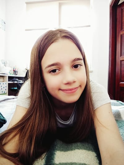 A young girl with long brown hair smiles at the camera, her face filling the foreground of the image. She is lying down, with her head resting on a striped blanket. The background shows a bedroom with a white wall, a desk with art supplies, and a wooden door. The lighting suggests it is daytime, possibly in the afternoon, with light streaming in from a window. The room appears to be in a residential setting in Radzymin, Poland.