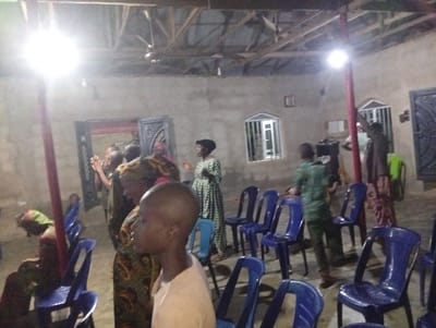 Inside a sparsely furnished building, a group gathers, illuminated by bright lights hanging from the ceiling beams. The room has simple concrete walls with framed windows. Several blue plastic chairs are arranged, some occupied. Individuals, predominantly women, stand facing an unseen focal point. Most are dressed in patterned fabrics and head coverings. A young man with a shaved head stands in the foreground, adding a sense of closeness to the scene.