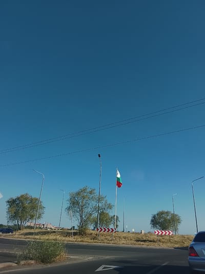 This media file captures a daytime scene in Aheloy, Bulgaria. The sky is a clear, deep blue, suggesting fair weather. The main subject is a Bulgarian flag, prominently displayed on a flagpole. The flag, with its white, green, and red horizontal stripes, is waving in the breeze.

The scene is set along a road, with asphalt visible in the foreground, indicating a transportation route. Several streetlights are positioned along the roadside, characteristic of urban or suburban areas. In the background, there are green trees with dry, brown grass on the elevated terrain, which might indicate a drier climate or season. Further in the distance, a cluster of buildings with light-colored facades is visible, suggesting a residential or commercial area.

Other objects in the scene include several cars, one of which is a silver sedan in the lower right corner of the frame, and another dark-colored car on the left. There are also directional road signs with red and white chevron patterns, which are commonly used to indicate curves or hazards on a road. Power lines are strung across the sky.

The image appears to be taken from a moving vehicle, given the angle and perspective. There is no discernible activity or event taking place; it is a static view of the environment. The time of day is clearly daytime, and the clear sky and bright lighting suggest it is likely midday or early afternoon. No people are visible in the frame. The overall impression is one of a roadside view in a semi-arid or transitional landscape under a bright, clear sky.