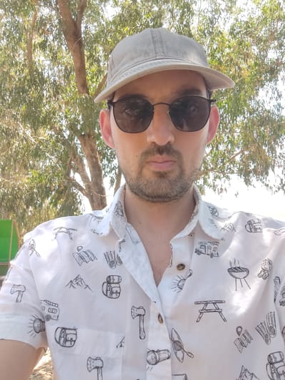 A man wearing a grey baseball cap, sunglasses, and a white short-sleeved shirt with black outdoor adventure illustrations is pictured from the chest up. He is outdoors, with a tree and foliage in the background. The location is likely within the Yoav Regional Council, Israel, based on the vegetation and arid appearance of the environment.