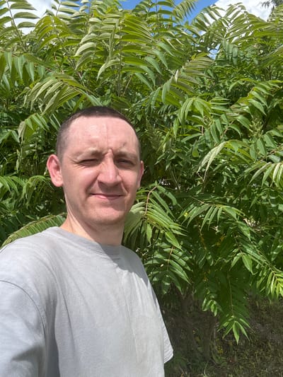 A man is taking a selfie in front of a large, leafy green tree. The man is wearing a grey t-shirt and has short, dark hair. He has a light complexion and appears to be in his 30s or 40s. The tree has compound leaves with many leaflets. The background shows a blue sky with some white clouds. The scene appears to be outdoors on a sunny day. The image was taken in Mazalava village, Vitebsk region, Belarus.
