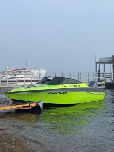 A neon green and black motorboat is docked at a wooden pier on a calm body of water. The boat has a registration number "E-210142" and the name "АНАКОНДА" printed on its side. In the background, another white passenger boat is docked at a pier with a sign that reads "ПРОГУЛ С ПРОЗР". The environment appears overcast with a light fog. The scene is in Listvyanka, Russia, a settlement on the northwestern shore of Lake Baikal. There are no people visible in the image.