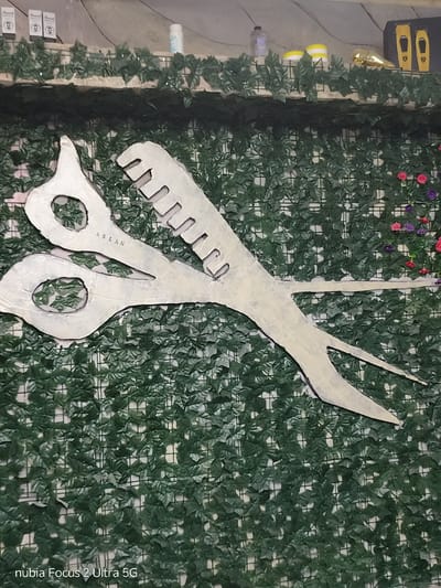 The image displays a large, stylized, silver pair of scissors with a comb attachment, mounted on a wall covered with artificial green ivy. The scissors appear to be a decorative element, possibly for a barbershop or salon. The background shows a shelf above the ivy wall, with various items including bottles, a golden object, and a black box with two yellow shapes. Some small pink and purple artificial flowers are visible amongst the ivy to the right. Text at the bottom of the image reads "nubia Focus 2 Ultra 5G". The scene is indoors, with a textured wall and artificial greenery.