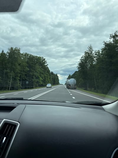 The image captures a view from the interior of a car, looking out onto a highway in the vicinity of Акцябрскі сельскі Савет, Belarus. The road ahead is paved, with clear white lane markings. On the left side of the road, a dense line of trees, primarily deciduous with some birch trees, borders the highway. A silver sedan is visible on the left lane. On the right side of the highway, a large tanker truck is traveling in the right lane, also surrounded by trees. The sky above is overcast with a thick layer of gray clouds. The dashboard of the car, including an air vent, is visible in the foreground.