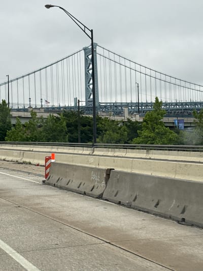 The image depicts a bridge structure with a suspension cable system visible. In the foreground, a concrete barrier runs along a roadway. A street lamp is mounted on a pole to the left, extending over the road. Further back, lush green trees are abundant. An American flag is displayed on a flagpole amidst the trees and bridge structure. The sky appears overcast, suggesting cloudy weather. The scene is likely outdoors, possibly on a highway or elevated road leading to or from the bridge. There is orange and white striped construction safety equipment on the road. The image's orientation is rotated, with the bridge appearing to stretch upwards. No people are visible in the frame. There is some graffiti on the concrete barrier in the foreground.