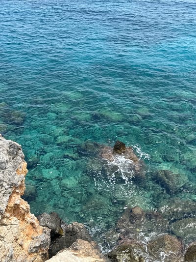 A high-angle shot captures the clear, turquoise waters of the Mediterranean Sea lapping against the rocky coastline of Rizokarpaso, Northern Cyprus. The water's transparency allows for a view of the submerged rocks and seabed, with gentle ripples disturbing the surface. In the foreground, rugged, textured rocks of varying shades of grey and orange form the edge of the land. The scene is devoid of any human presence or activity, presenting a natural and serene coastal landscape.