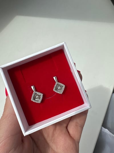 The image shows a pair of diamond earrings displayed in a white jewelry box lined with red velvet. The earrings are square-shaped with intricate diamond detailing. A hand with red nail polish is holding the jewelry box. The background is a plain white surface. The image does not provide any information or visual cues to determine its location in Lytkarino, Russia.
