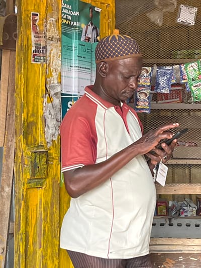 An elderly man wearing a patterned hat is absorbed in his phone. He's dressed in a two-tone shirt with red sleeves and a white torso. A charger is connected to his phone. He is leaning on a yellow counter plastered with posters. Behind him are shelves stocked with various packaged goods. The photo is taken at a slightly off-kilter angle.