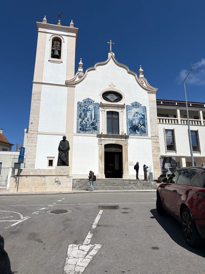 The image shows a street view of the Igreja do Carmo church in Aveiro, Portugal, with a reddish car parked on the right side of the frame. The church is white, adorned with blue and white tile panels, a bell tower, and a statue of João Evangelista de Lima Vidal. A few people are seen on the steps leading up to the church's entrance. The street shows pedestrian crossings and markings. A signpost is visible near the church's base. The sky is a clear blue. Adjacent buildings are visible behind the church.