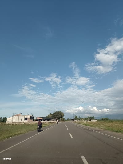 The image shows a road in Dayah Kruet, Indonesia. The sky is blue with scattered clouds. On the left side of the road, there is a building under construction and a field of grass. A person is riding a motorcycle on the road, heading away from the camera. On the right side of the road, there are some trees and a mountain in the distance. There are also some streetlights visible. The road has white lines marking the lanes. The overall scene depicts a rural or semi-rural setting on a sunny day.
