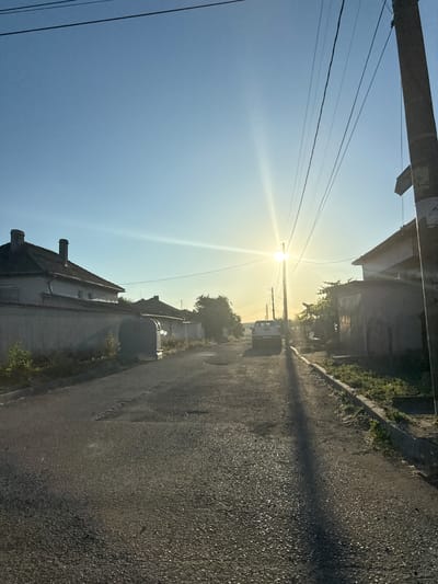 This is a vertical photograph of a street scene in Marinka, Bulgaria, at sunrise. The sun is low in the sky, casting a bright glare and long shadows. Power lines stretch across the frame from a utility pole on the left. On the right side of the street, a paved road runs parallel to a sidewalk lined with green vegetation. Buildings with tiled roofs and chimneys are visible on either side of the street. A white van is parked on the right side of the road. The overall mood is serene, with the warm light of dawn illuminating the quiet street. There is no visible text in the image.