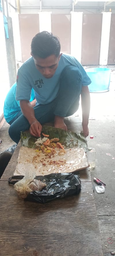 An individual is captured in a crouched position, actively engaging with a meal laid out on a wooden surface in Sulaeman, Indonesia. The male subject, wearing a light blue t-shirt with "UNIT 10" printed on it and blue jeans, is barefoot. His right hand is positioned over a banana leaf and white material containing rice, several orange-colored food items (possibly shrimp or fried snacks), and other mixed food components. A partially obscured second individual, also in a light blue t-shirt, is visible in the background to the left.

On the wooden surface, adjacent to the meal, are a clear plastic bottle with a red cap containing water, a small clear plastic bag holding brown, granular material, and a larger black plastic bag beneath it. To the right of the wooden surface, on a concrete ground, are two cigarette lighters (one red, one black/red), crumpled metallic packaging, and a red rubber band.

The immediate environment includes a dark brown wooden plank surface in the foreground and concrete ground to the right. The background features light brown vertical panels or doors, behind which a bright light source is visible. A light blue plastic tub is positioned on the concrete ground in the mid-background. Structural elements, including metal beams and piping, are also visible above and to the left in the background.